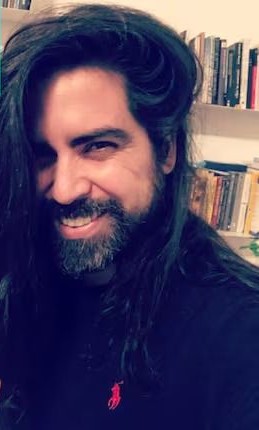 Nico Saraintaris sonriendo, con pelo largo y barba, frente a una biblioteca.